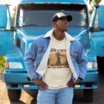 Man wearing Frontier Rogue Beaten and Battered But Still Going Strong graphic tee in front of blue semi truck