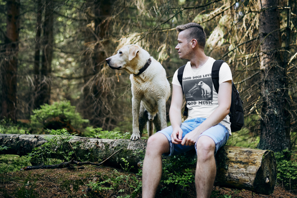 Man wearing Frontier Rogue Good Dogs Love Good Music graphic tee sitting on log with dog in outdoor setting