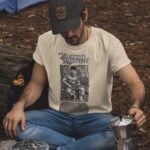Man wearing Frontier Rogue Theodore Roosevelt graphic tee sitting in woods with camping gear